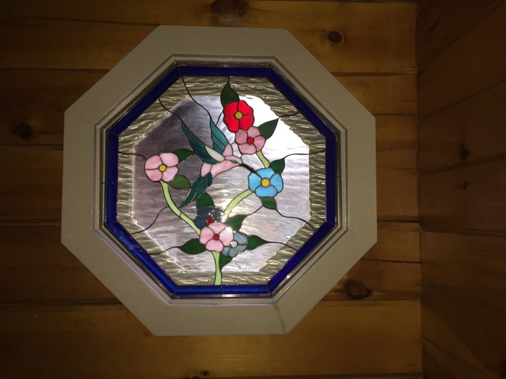 An octagonal stained glass panel depicting a pink hummingbird with green wings gracefully hovering among pink, red, and blue flowers with green leaves. The background is clear, textured glass, framed by a royal blue border. The panel is set within a light-colored octagonal frame against a rustic wooden plank wall.