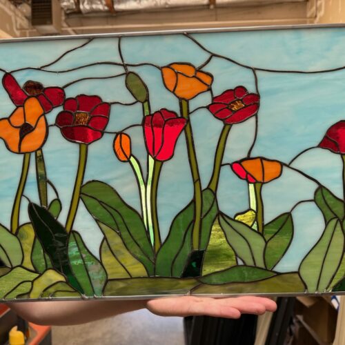 A rectangular leaded stained glass panel depicting a vibrant field of poppy flowers. The poppies are in shades of bright red and orange with varied green leaves and stems, all set against a wispy light blue sky.
