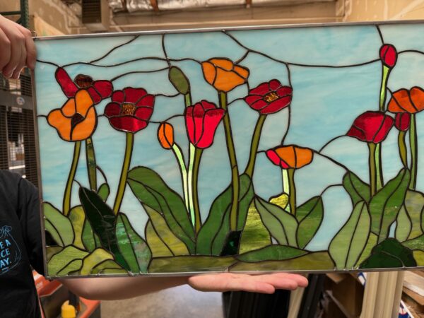 A rectangular leaded stained glass panel depicting a vibrant field of poppy flowers. The poppies are in shades of bright red and orange with varied green leaves and stems, all set against a wispy light blue sky.