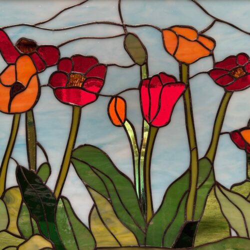 A rectangular leaded stained glass panel depicting a vibrant field of poppy flowers. The poppies are in shades of bright red and orange with varied green leaves and stems, all set against a wispy light blue sky.