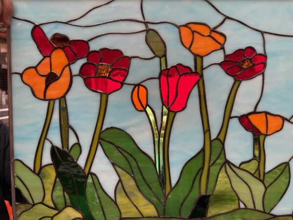 A rectangular leaded stained glass panel depicting a vibrant field of poppy flowers. The poppies are in shades of bright red and orange with varied green leaves and stems, all set against a wispy light blue sky.