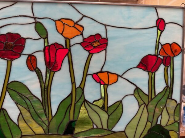A rectangular leaded stained glass panel depicting a vibrant field of poppy flowers. The poppies are in shades of bright red and orange with varied green leaves and stems, all set against a wispy light blue sky.