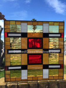 A square stained glass panel with a bold, modern geometric design featuring a prominent central red square, surrounded by amber, yellow, and white rectangular glass pieces, all joined by black lead lines. The panel is held outdoors against a blue sky.