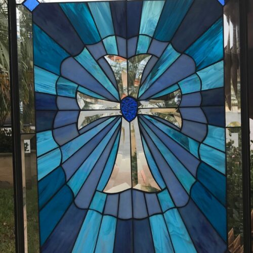 A rectangular stained glass panel featuring a prominent clear beveled glass cross at its center. The cross is surrounded by a sunburst pattern of variegated blue stained glass, including shades of navy, medium blue, and turquoise. The outer edges have clear glass borders with small square blue glass accents in the top corners. The panel has a dark metal frame and is seen against an outdoor backdrop.