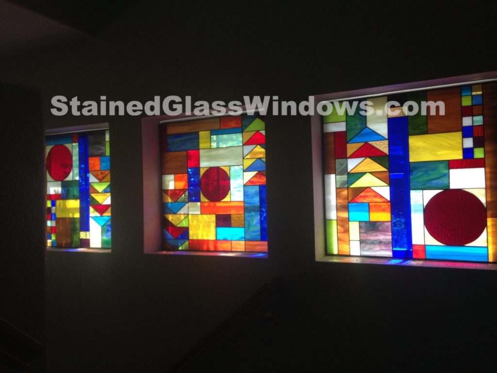 Three rectangular stained glass window panels in a dark interior wall, featuring a vibrant, abstract geometric design with prominent red circles, various sized rectangles, and triangles in shades of blue, yellow, red, orange, and clear glass.