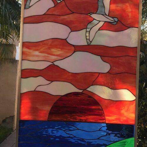 A tall, rectangular leaded stained glass art panel depicting a vibrant coastal sunset. The top shows an orange and red sky with white clouds, and two grey and white seagulls in flight. Below, a large red sun sets over a deep blue ocean with a frothy white wave. The bottom third features rolling green hills with a foreground of red and light-colored poppy flowers.