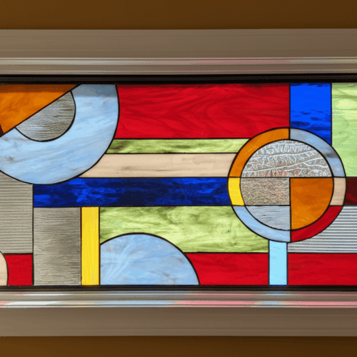 A modern, abstract stained glass window with a geometric design. The asymmetrical composition features overlapping circles, semi-circles, and rectangles made from a mix of textured glass and bold colored glass in shades of red, blue, yellow, and green.