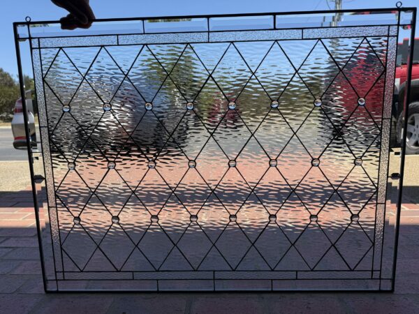 A rectangular clear stained glass panel with a diamond lattice pattern, featuring round clear jewels at intersections. The panel includes textured clear glass and beveled clear glass elements along its borders, all joined by black lead lines. It is held up outdoors, reflecting the surrounding environment.