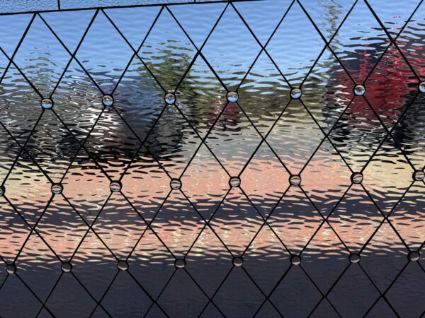 A clear leaded stained glass panel featuring a diamond grid pattern of heavily textured, rippled glass. Small, clear glass jewels are embedded at each intersection, distorting the reflections of blue, green, red, and orange seen through the glass.