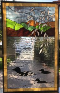 A rectangular stained glass panel depicting a serene lake landscape. In the foreground, three loons swim on clear, rippled textured glass water, with one large loon featuring prominent black and white patterns and two smaller dark loons. Above the water are rolling hills in shades of green and brown textured glass, set beneath a soft blue and gray sky. Delicate white, elongated leaves hang from the top right, adding depth to the natural scene. The entire panel is framed by a golden-yellow border.