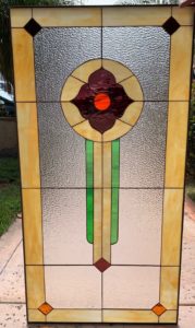 A rectangular leaded stained glass panel featuring a central circular floral motif with dark burgundy petals and a bright orange center, set against clear textured glass. The panel has a wide amber-yellow border with dark brown diamonds in the top corners and orange diamonds in the bottom corners. Two vertical green strips and a cream strip extend downwards from the central design.