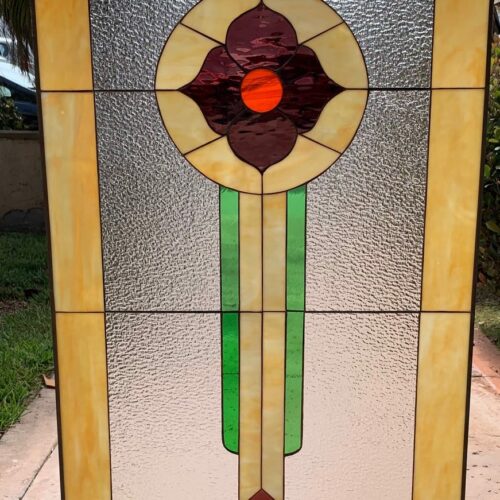 A rectangular leaded stained glass panel featuring a central circular floral motif with dark burgundy petals and a bright orange center, set against clear textured glass. The panel has a wide amber-yellow border with dark brown diamonds in the top corners and orange diamonds in the bottom corners. Two vertical green strips and a cream strip extend downwards from the central design.
