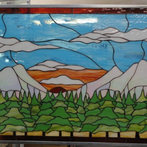 A rectangular leaded stained glass panel depicting a vibrant mountain landscape. The scene features light-colored, snow-capped mountains in the mid-ground, a dense evergreen forest with varying shades of green fir trees in the foreground, and an orange and red sunset or sunrise over a distant horizon. The sky is a bright blue with wispy white clouds, all framed by a narrow border of deep reddish-brown glass.