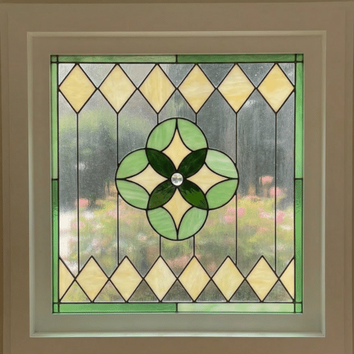 A square, leaded stained glass panel with a central clover design in various shades of green glass and a clear faceted jewel. The design is framed by light yellow diamond shapes and clear textured glass, bordered by solid green glass.