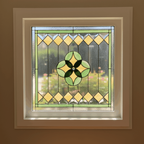 A square, vinyl-framed insulated stained glass window with a geometric design. The center features a four-leaf clover medallion in light and dark green glass, surrounded by a border of pale yellow diamond-shaped glass pieces and clear textured glass.