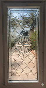 A rectangular, leaded stained glass window panel featuring a prominent clear beveled compass rose medallion at its center, surrounded by clear textured glass in a diamond pattern, set in a white frame.