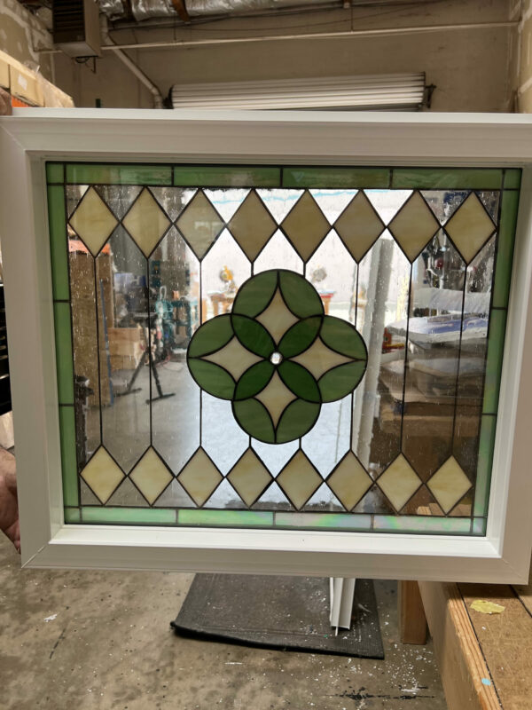 A square, vinyl-framed insulated stained glass window with a geometric design. The center features a four-leaf clover medallion in light and dark green glass, surrounded by a border of pale yellow diamond-shaped glass pieces and clear textured glass.