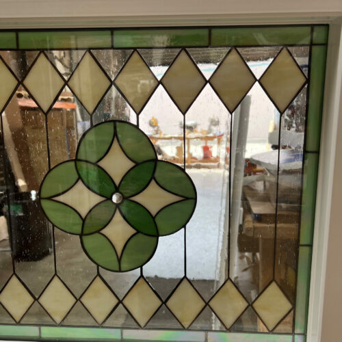 A square, vinyl-framed insulated stained glass window with a geometric design. The center features a four-leaf clover medallion in light and dark green glass, surrounded by a border of pale yellow diamond-shaped glass pieces and clear textured glass.