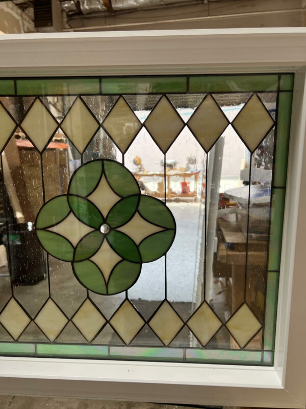 A square, vinyl-framed insulated stained glass window with a geometric design. The center features a four-leaf clover medallion in light and dark green glass, surrounded by a border of pale yellow diamond-shaped glass pieces and clear textured glass.