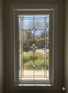 A rectangular clear stained glass window panel with a symmetrical geometric pattern made from various textured glass pieces and small faceted bevels, joined by dark lead lines. The panel offers privacy while allowing light to filter through, showcasing a vibrant green garden beyond.