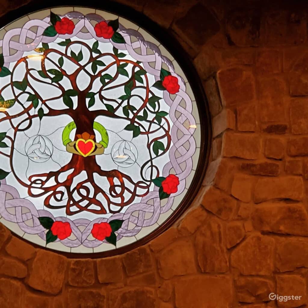 A circular stained glass window featuring a vibrant Celtic Tree of Life design with brown roots and trunk, green leaves, and a gold Claddagh ring with a red heart at its base. The clear glass background is bordered by intricate light purple Celtic knotwork interwoven with red roses and green leaves. The window is set into a rustic stone wall.
