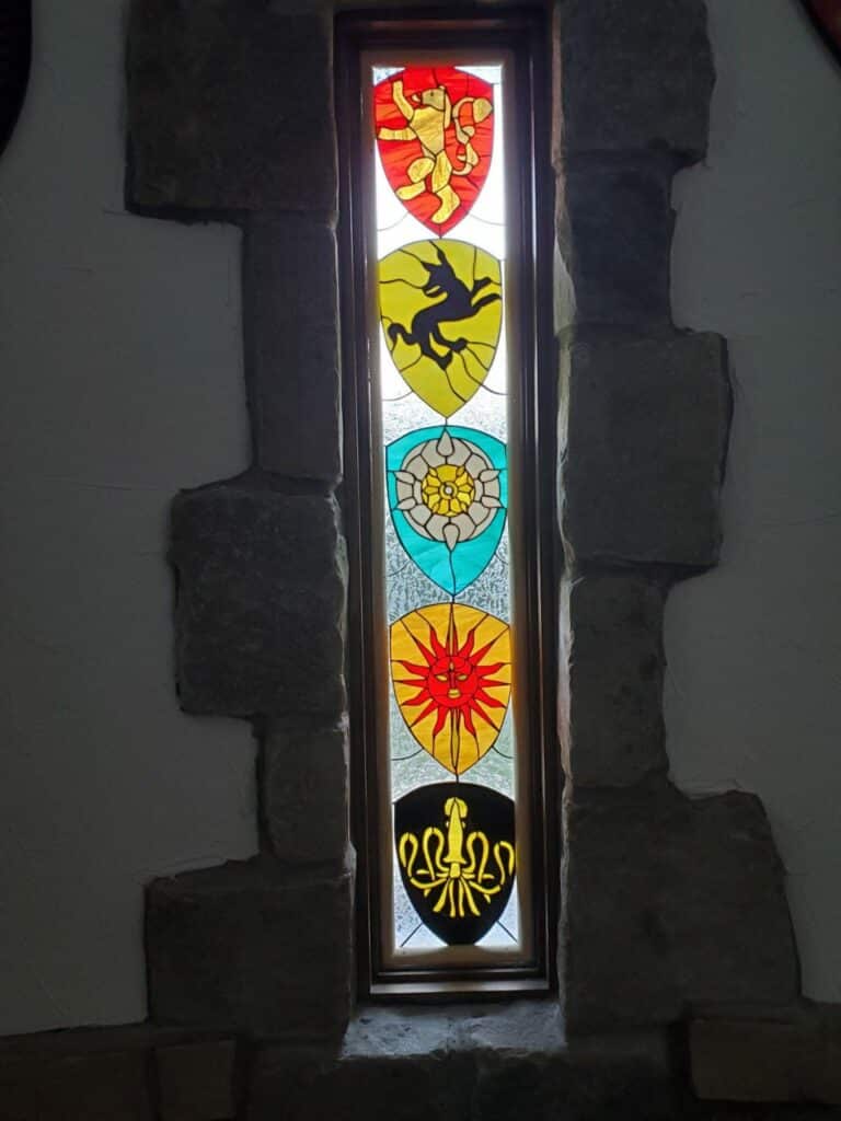 A tall, narrow, leaded stained glass window embedded in a textured stone wall. It features five vibrant shield-shaped panels stacked vertically, each depicting a distinct emblem: a golden heraldic lion on a red field, a black unicorn-like creature on a yellow field, a white and yellow Tudor rose on a teal field, a stylized red sun with a face on a yellow field, and a golden octopus or squid on a black field.