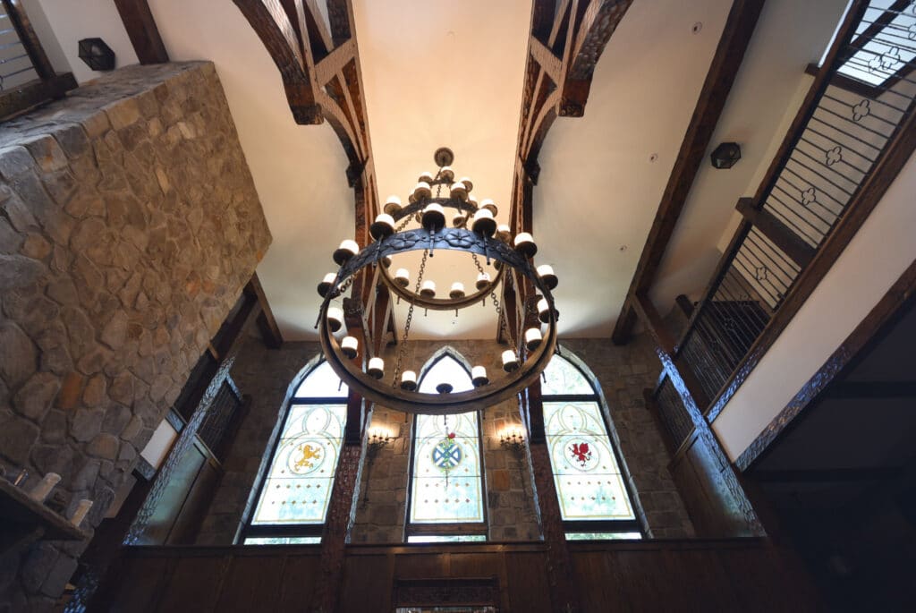 Three tall, arched stained glass windows set into a stone wall in a grand, high-ceilinged room with dark wood beams and a large wrought-iron chandelier. The windows feature clear and light-colored glass with central heraldic emblems: a yellow lion rampant, a white cross with red thistles, and a red Welsh dragon. Geometric patterns surround the emblems. A second-story balcony with an iron railing is visible on the right.