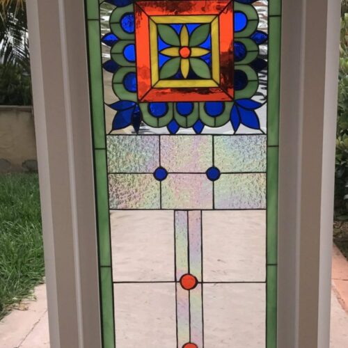 A rectangular stained glass door panel, framed in white, featuring a vivid geometric design. It has a bright green border, large sections of clear textured glass, and a central square medallion. The medallion is outlined in orange, with a yellow and green floral center, surrounded by deep royal blue leaf-like shapes. Small circular blue and red glass accents are placed throughout the clear sections.