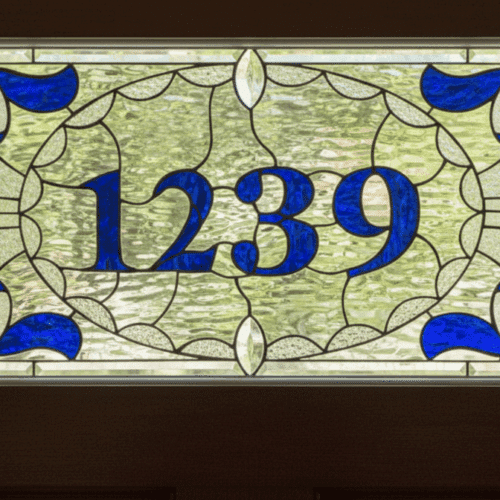A rectangular stained glass transom window displaying the address '1239'. The numbers are in a bold, blue font, centered within an ornate oval frame of white and blue scrollwork. The background is a textured, clear privacy glass.