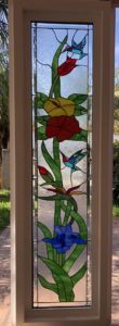 A tall, rectangular leaded stained glass panel depicting a vibrant tropical scene. Two hummingbirds with blue, red, and teal plumage are shown amidst colorful flowers including red, yellow, and blue blooms, and lush green foliage. The background is clear, textured glass, providing privacy.