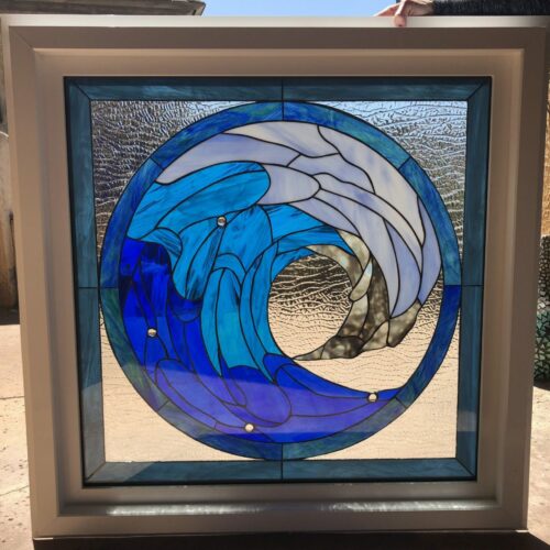 A square, white-framed stained glass window panel featuring a circular design of a dynamic ocean wave. The wave is rendered in various shades of blue and white textured and opaque glass, with clear heavily textured glass surrounding it and light sandy-toned textured glass at its base. Black lead lines define the intricate shapes, and clear round glass jewels are embedded in the blue sections, catching the light.