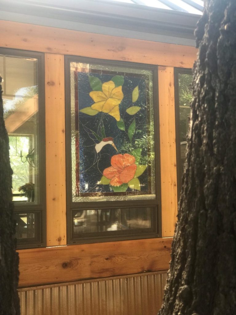 A rectangular stained glass window panel depicting a white and red hummingbird hovering between a large yellow flower and a large orange flower, surrounded by green leaves on a dark blue textured background. The panel is installed in a window unit framed by light wooden beams.