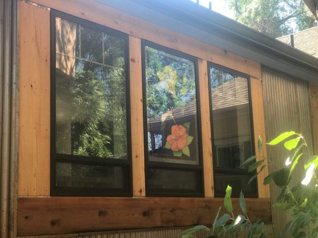 An exterior view of three rectangular windows set into a building with a rustic facade of natural wood and corrugated metal. The central window features a custom stained glass panel with a large, vibrant orange-red hibiscus flower, green leaves, a smaller yellow flower, and a white hummingbird. Green trees and sunlight reflect in the glass.