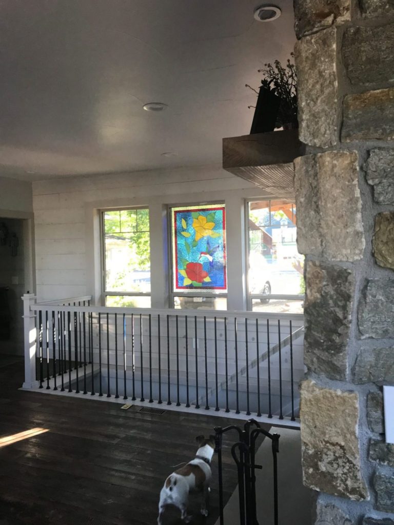 A rectangular stained glass panel depicting a vibrant hummingbird flying among red and yellow flowers with green foliage, set against a bright blue background, installed in a home window above a white and black railing.