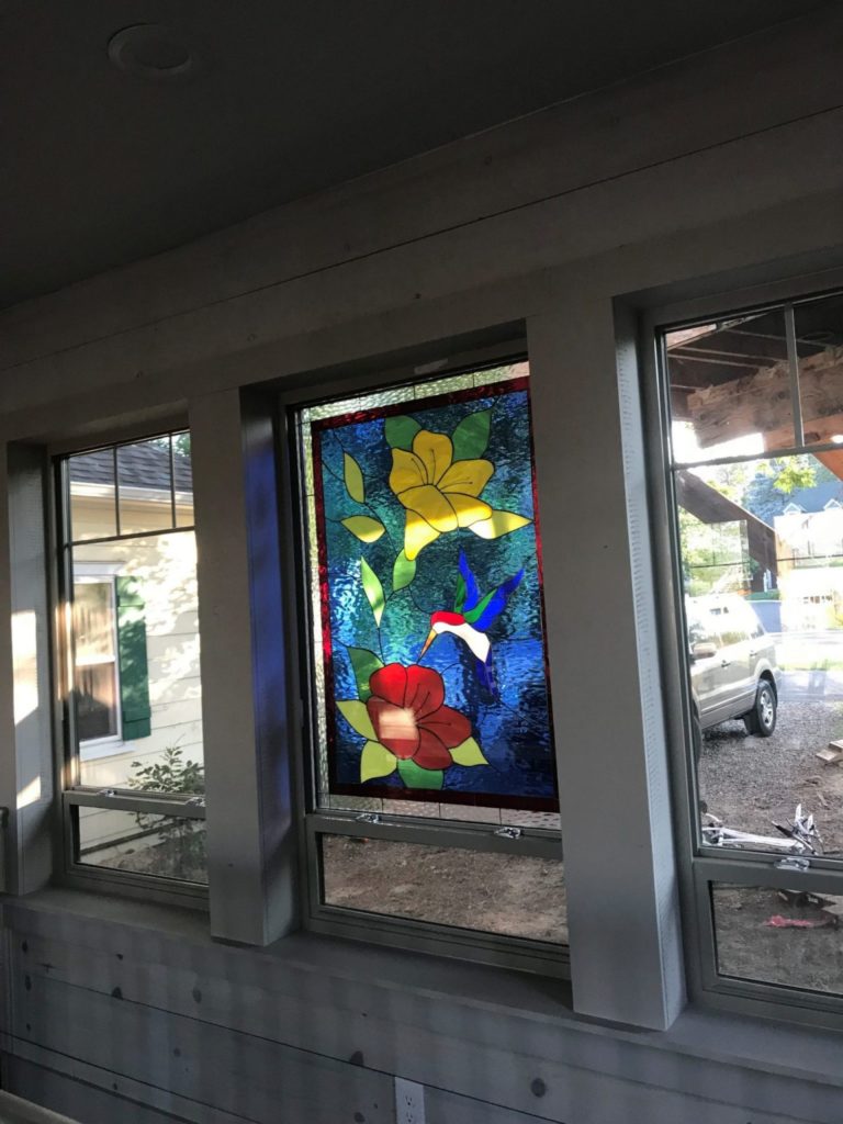 A rectangular stained glass window panel depicting a vibrant hummingbird with red, white, and blue plumage, hovering between a large red flower and a large yellow flower. Green leaves surround the flowers, all set against a deep, textured blue background.