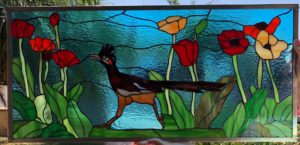 A rectangular leaded stained glass panel depicting a vibrant desert scene with a brown roadrunner bird walking among red and orange poppies and lush green foliage, set against a textured blue sky.