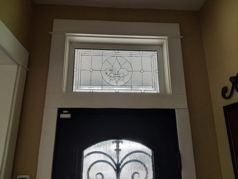 A rectangular, clear leaded stained glass transom window with a central beveled Fleur-de-Lis design, surrounded by textured and smooth clear glass panels, installed above a dark front door.