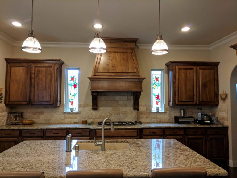 Two tall, rectangular stained glass window panels, each depicting red roses and green and yellow leaves on a clear background, set into a kitchen with wooden cabinets and a granite island.