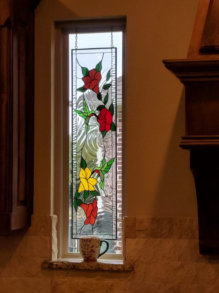 A rectangular, leaded stained glass panel with a nature-themed design. It shows two red and one yellow flower with green leaves, and two green hummingbirds with red accents, all against a clear, swirled textured glass background, hanging in a vertical window.