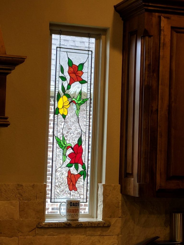 A vertical leaded stained glass window panel featuring vibrant red and yellow flowers with green leaves and two hummingbirds, set against a clear textured background, hanging in a narrow window above a kitchen counter.