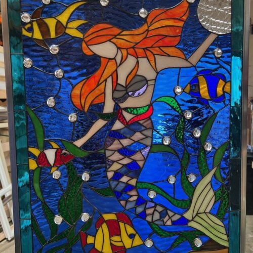 A vibrant rectangular stained glass panel featuring a red-haired mermaid with a scaled tail amidst an ocean scene. Colorful fish, dark green seaweed, and clear round glass jewels resembling bubbles are set against a rich blue textured water background. The mermaid holds a silver, textured disc.