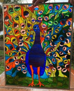 A rectangular, brightly colored leaded stained glass panel depicting a majestic peacock with its tail feathers fully fanned out. The peacock's body is a rich dark blue, with a blue head and bright orange beak and legs. Its elaborate tail feathers explode in a mosaic of vibrant blues, greens, yellows, and oranges, with distinct eye-like patterns. The background suggests green foliage. The entire panel is encased in a slim metal frame.