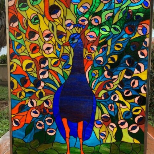 A rectangular, brightly colored leaded stained glass panel depicting a majestic peacock with its tail feathers fully fanned out. The peacock's body is a rich dark blue, with a blue head and bright orange beak and legs. Its elaborate tail feathers explode in a mosaic of vibrant blues, greens, yellows, and oranges, with distinct eye-like patterns. The background suggests green foliage. The entire panel is encased in a slim metal frame.