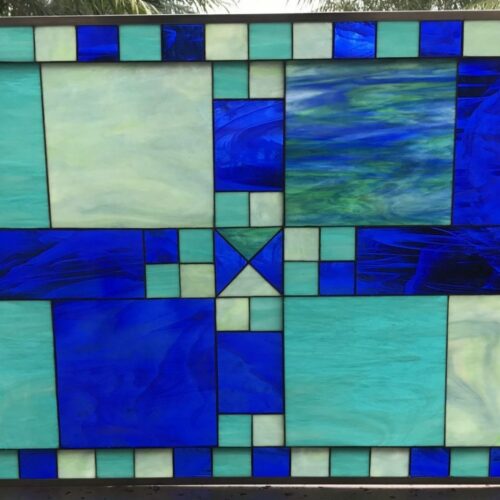 A rectangular leaded stained glass panel featuring an abstract geometric design composed of various shades of blue, aqua, and pale green glass. The design showcases large rectangular and square sections with dynamic, swirled blue glass, complemented by smoother aqua and pale green pieces, all framed by a border of alternating small squares and geometric cross patterns.