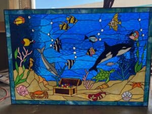 A rectangular leaded stained glass panel depicting a lively underwater ocean scene. The vibrant blue water, featuring clear glass bubbles, is filled with marine life including a large black and white orca, a gray dolphin, a green sea turtle, numerous colorful tropical fish (e.g., angelfish, clownfish), a seahorse, crab, and lobster. The sandy seafloor showcases an open treasure chest filled with colorful gems, various seashells, a starfish, and coral. The panel is framed in a border of green and blue glass.