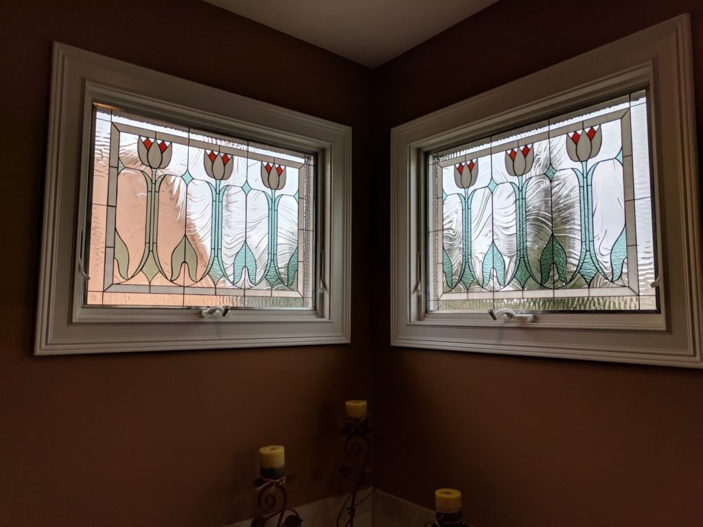 Two rectangular custom leaded stained glass window panels featuring a repeating Art Nouveau-inspired design with stylized white tulips, red-orange centers, green stems, and flowing clear textured glass. The panels are installed in white window frames in a corner room with brown walls.