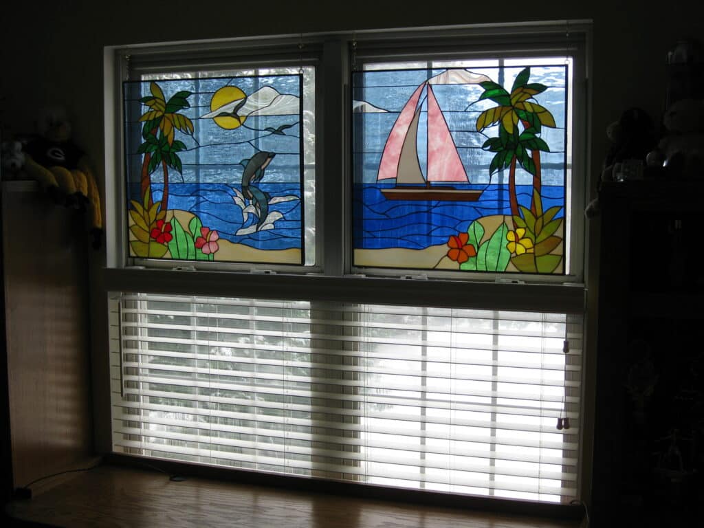 A pair of rectangular stained glass window panels installed in the upper part of a window. The left panel depicts a tropical scene with a blue dolphin leaping from ocean waves, green palm trees on a small sandy island with red and yellow flowers, a yellow sun, and white clouds in a light blue sky. The right panel shows another tropical scene with a white sailboat with a prominent pink sail floating on blue ocean waves, green palm trees on a small sandy island with orange and yellow flowers, and white clouds in a light blue sky.