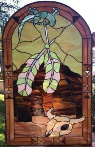 An arched leaded stained glass panel in a Southwestern style, featuring a green gecko at the top, two pale green and pink stylized feathers in the center, and a cream steer skull at the bottom. The background glass is golden yellow and streaky amber-brown, framed by a thick geometric amber border with small blue and black accent triangles.