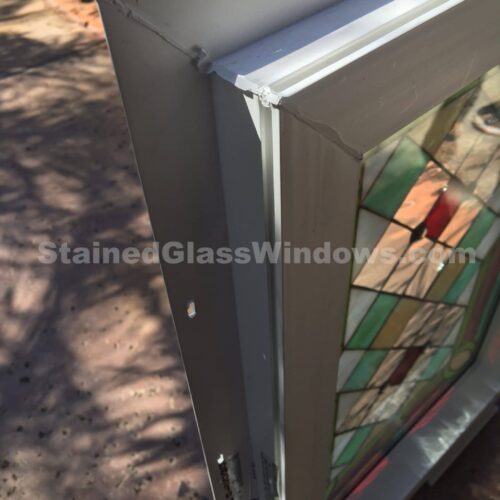 A close-up, angled view of a custom-framed insulated stained glass window unit. The white vinyl or aluminum frame shows pre-drilled holes for installation. The stained glass within features a vibrant geometric diamond and rectangular pattern in shades of green, amber, and red, interspersed with clear textured glass. Sunlight illuminates the unit, casting shadows on the ground.