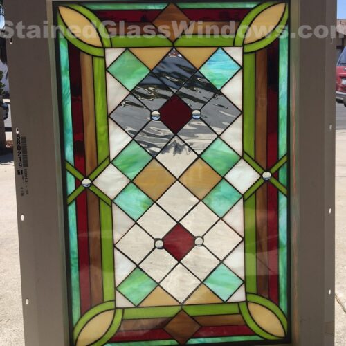 A rectangular leaded stained glass panel in a dark bronze frame, featuring a vibrant geometric diamond grid design. The panel combines rich ruby red, various shades of green, warm amber, opalescent white, and clear textured glass, with beveled elements and sparkling clear round glass jewels. The design evokes classic Prairie and Art Deco styles.